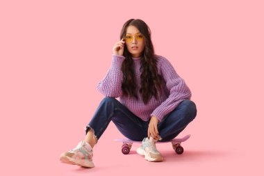 Pembe arka planda kaykayın üzerinde oturan şık kıyafetli ve modern kulaklıklı güzel bir Asyalı kadın.