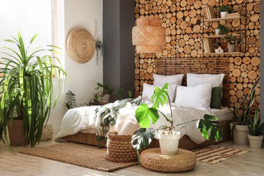 Yatak odasının içinde yeşil bitkiler, kitaplık ve puf var.