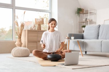 Evde yoga minderinde glukofon çalan dizüstü bilgisayarlı genç bir kadın.