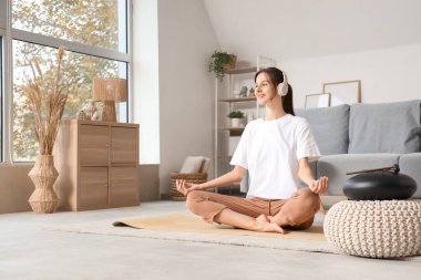 Kulaklıklı genç bir kadın evde yoga minderi üzerinde meditasyon yapıyor.