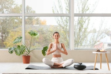 Evde pencerenin yanında meditasyon yapan genç bir kadın.