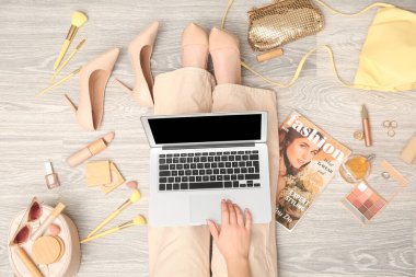 Modern dizüstü bilgisayarı, farklı dekoratif kozmetik ürünleri, aksesuarları ve dergisi olan bir kadın açık tahta zeminde oturuyor. Çevrimiçi alışveriş kavramı