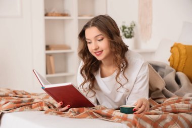 Evdeki yatağında bir fincan kahve içip kitap okuyan güzel, genç bir kadın.