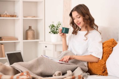 Güzel, genç, mutlu bir kadın. Bir fincan kahve ve dizüstü bilgisayarla evde kitap okuyor.