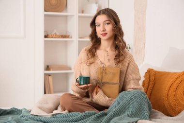Güzel, genç, mutlu bir kadın, elinde kahve ve kitaplarla evde, yatağında oturuyor.