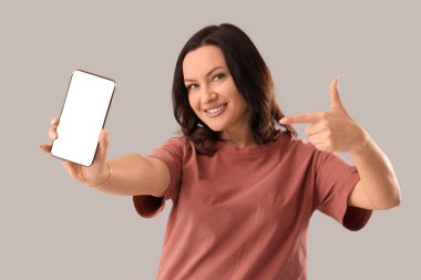 Gri arka planda cep telefonuna işaret eden olgun mutlu kadın.