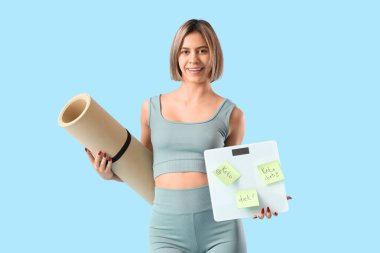 Mavi arka planda KETO DIET ve fitness paspası ile ağırlık tartıları tutan sportif genç kadın