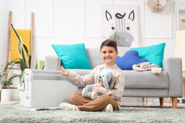 Cute boy with toy bear and electric convector heater sitting on carpet at home clipart