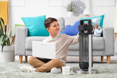 Evde elektrikli ısıtıcının yanında oturan dizüstü bilgisayarı olan sevimli çocuk.