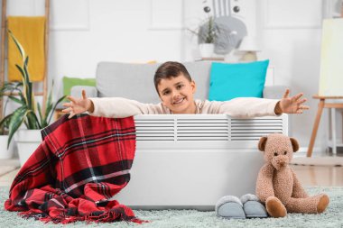 Cute boy with electric convector heater and toy bear at home clipart