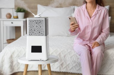 Yatak odasında cep telefonu kullanan kadınlara karşı modern hava nemlendirici, yakın plan.