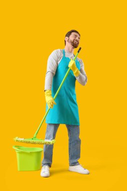 Sarı arka planda paspas ve kova ile şarkı söyleyen erkek hademe.