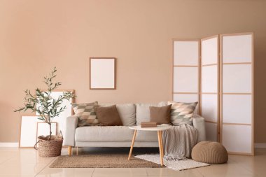 Modern oturma odasının içinde gri kanepe, masa, zeytin ağacı ve bej duvarda katlanır ekran.