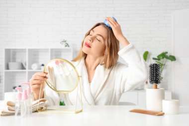 Evde aynalı genç bir kadın saç derisi masaj aleti kullanıyor.