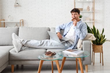 Yakışıklı genç adam, modern bilgisayarla çikolata yiyor ve oturma odasında kanepede oturuyor.