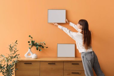 Evdeki turuncu duvara boş bir çerçeve asan genç bir kadın.