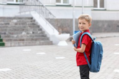 Sırt çantalı ve okulun yanında kitapları olan sevimli bir çocuk.