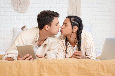 Yatak odasında bilgisayarı ve laptopu olan genç çift öpüşüyor.