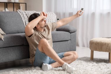 Yakışıklı genç adam oturma odasında klimalı uzaktan kumandayla yerde oturuyor.