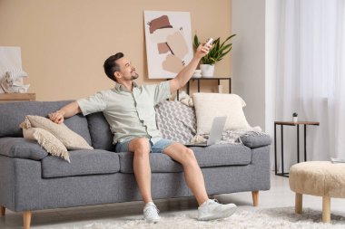 Genç adam oturma odasındaki bej duvarın yanındaki kanepede otururken klimayı açıyor.