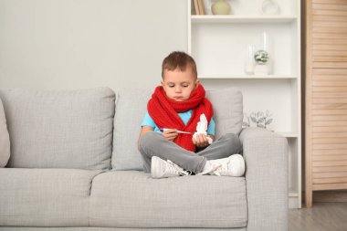 Evdeki kanepede oturan termometresi ve mendili olan hasta çocuk.