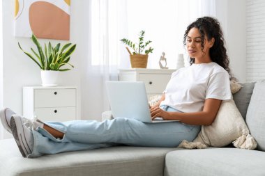 Evdeki kanepede dizüstü bilgisayarı olan güzel bir Afro-Amerikalı kadın.