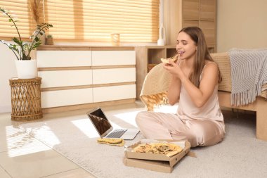 Pijamalı, dizüstü bilgisayarlı, pizza yiyen ve evinde yerde oturan genç bir kadın.
