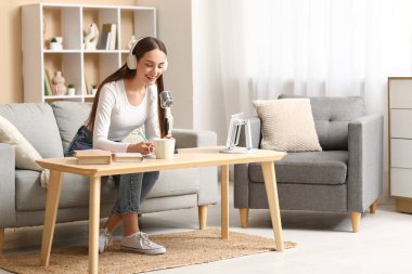 Kulaklık takan, elinde mikrofon ve not defteri olan genç bir kadın evde podcast yapıyor.