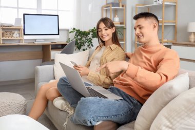 Genç çift evdeki koltukta dizüstü bilgisayar ve cep telefonu kullanıyor.