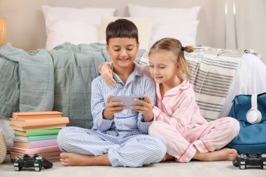Cep telefonları ve kitaplarıyla evde video oyunları oynayan pijamalı şirin çocuklar.