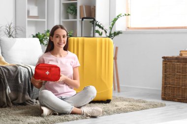 Evinde yerde oturan bavulu ve ilk yardım çantasıyla mutlu genç bir kadın. Seyahat kavramı