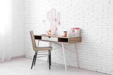 Ofisteki beyaz tuğla duvarın yanında sandalye, lamba ve kırtasiye malzemesi olan şık bir iş yeri.
