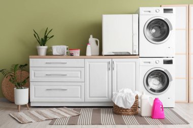 Çamaşır makineleri, deterjan şişeleri ve hasır sepeti olan modern çamaşır odası.