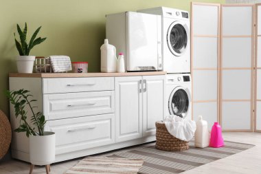 Çamaşır makineleri, deterjan şişeleri ve hasır sepeti olan modern çamaşır odası.