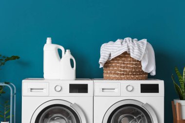 Çamaşır makinesi ve deterjan şişeleri mavi duvarın yanında çamaşır sepeti.