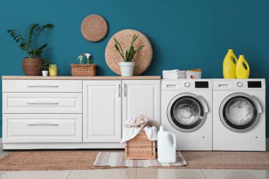 Çamaşır makineleri, deterjan şişeleri ve hasır sepeti olan modern çamaşır odası.