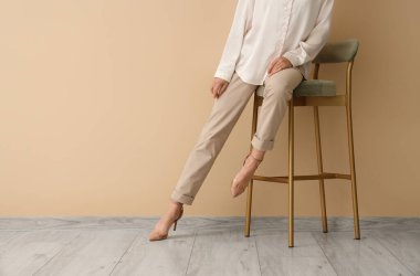 Topuklu giyen zarif kadın bej duvarın yanındaki taburede oturuyor.