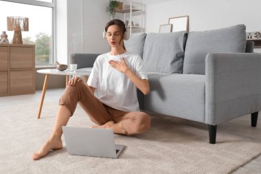 Kulaklıklı genç bir kadın evde laptopuyla meditasyon yapıyor.