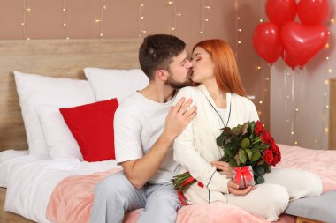 Pijamalı, gül demeti ve hediye kutusuyla evdeki yatakta oturan genç mutlu çift. Sevgililer Günü kutlaması