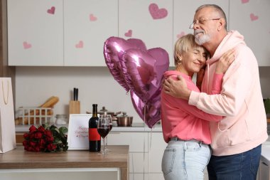 Happy mature couple with balloons, glasses of wine and roses bouquet hugging in kitchen at home. Valentine's Day celebration