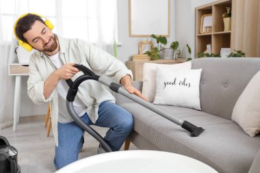 Kulaklıklı yakışıklı adam evdeki elektrikli süpürge kanepesini temizliyor.