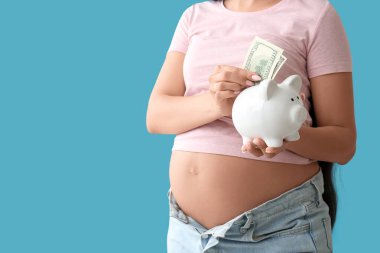 Genç hamile bir kadın mavi arka plandaki kumbaraya para yatırıyor, yakın plan. Annelik Faydası konsepti