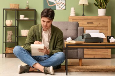 Oturma odasında yerde oturan dizüstü bilgisayarı ve klasik daktilosu olan genç bir yazar.