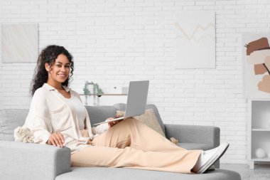 Evdeki kanepede dizüstü bilgisayarı olan güzel bir Afro-Amerikalı kadın.