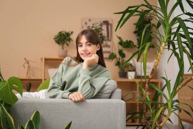 Oturma odasındaki çiçeklerin yanındaki kanepede dinlenen genç bir kadın.