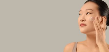 Genç Asyalı kadın, gri arkaplan üzerine yüz kremi sürüyor.