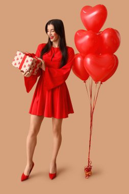 Yüzünde öpücük izleri olan güzel bir genç kadın. Elinde hediye kutusu ve kahverengi arka planda kalp şeklinde balonlar var.