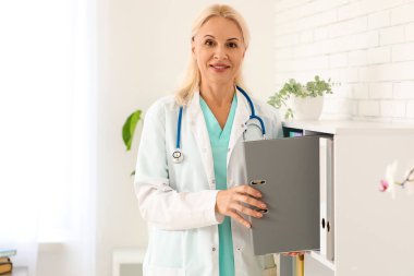 Olgun kadın doktor klinikteki raftan klasör alıyor.