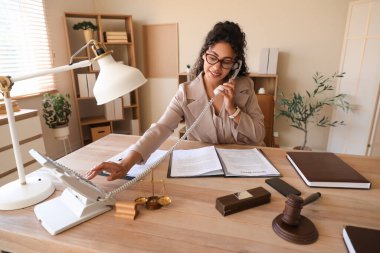 Female African-American lawyer talking by telephone at table in office clipart