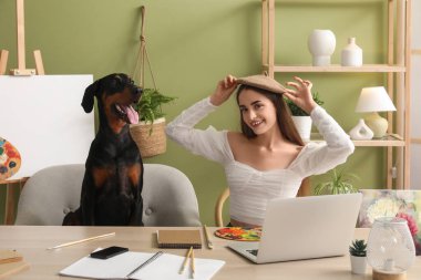 Tatlı Doberman köpeği ve dizüstü bilgisayarı olan güzel, mutlu, genç bir kadın. Evdeki şövalenin yanında palet boyuyor.